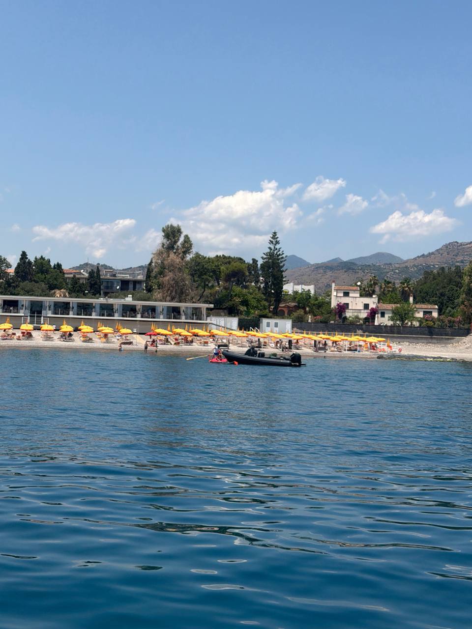 Beach umbrellas, tender, calm blue water, and Sicilian hillside at the Taormina swim stop